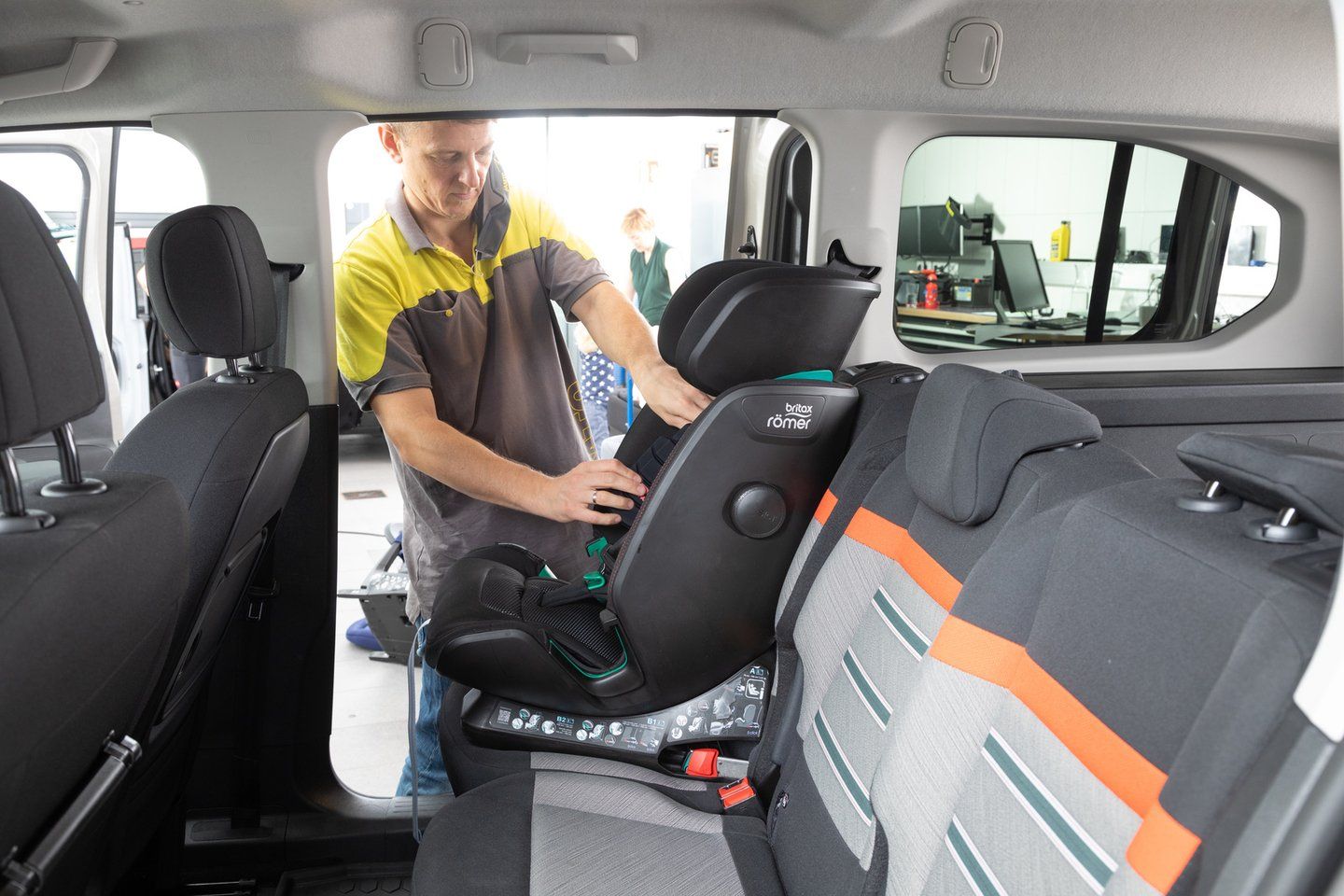 Un technicien en polo jaune et gris installe un siège auto Britax Römer noir sur la banquette arrière d'un ludospace, vu depuis l'intérieur du véhicule