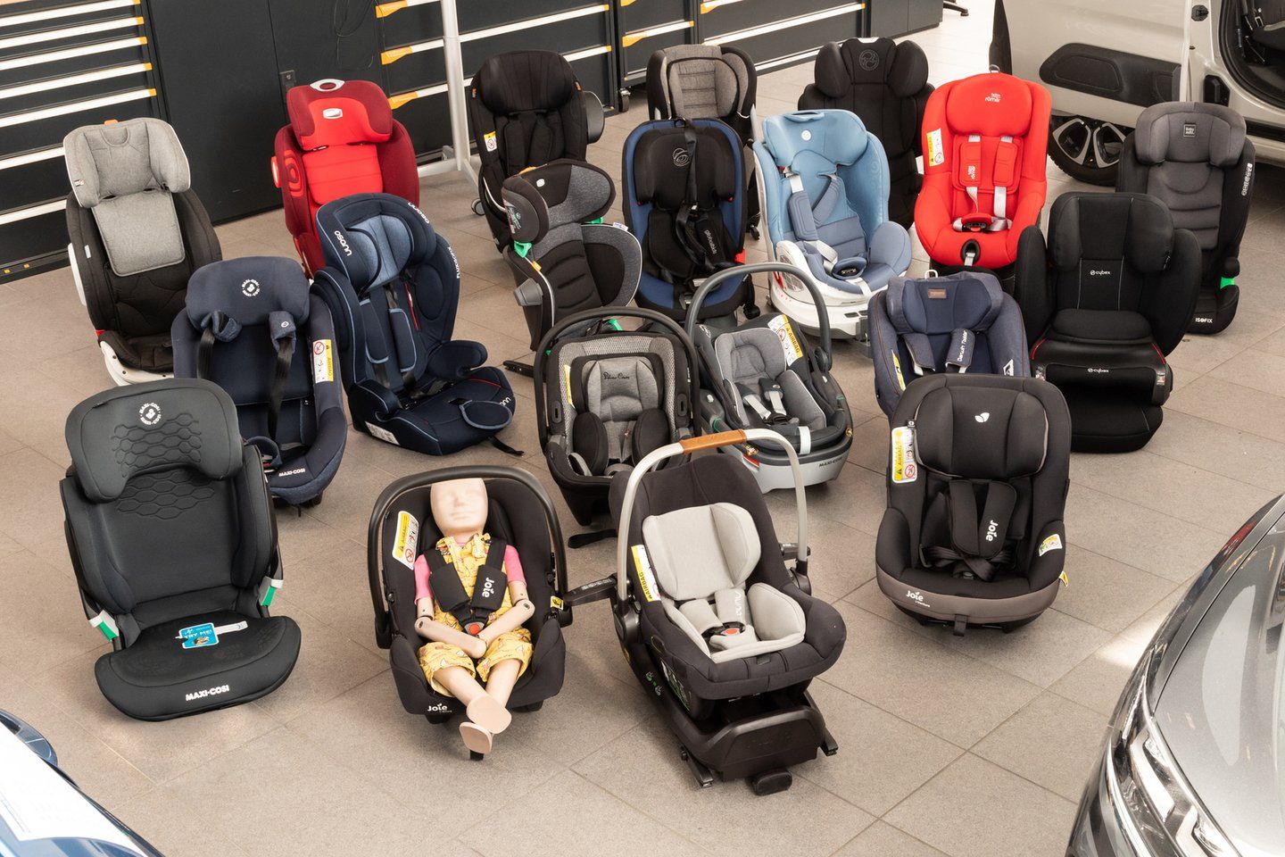 Une vingtaine de sièges auto et coques bébé du test disposés sur le sol d'un atelier, l'une des coques occupée par une poupée-mannequin