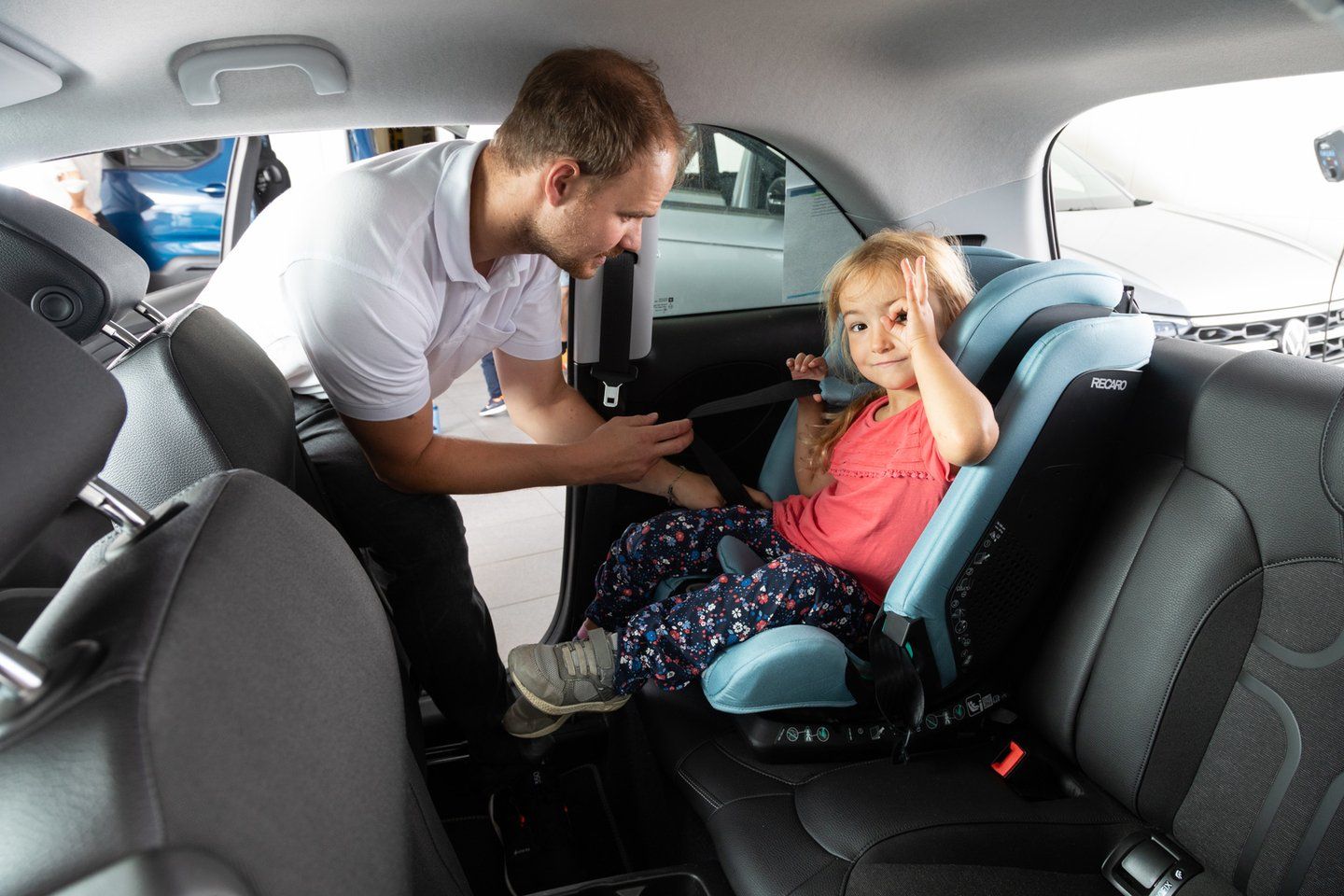 Un adulte en polo blanc tend la ceinture du véhicule vers une fillette assise dans un siège auto Recaro bleu clair, qui fait un signe « OK » avec la main