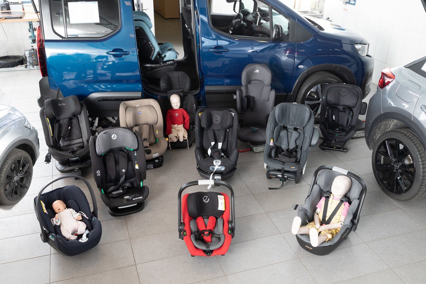 Sièges auto et coques bébé du test regroupés sur un sol carrelé devant un ludospace bleu à portière ouverte, certains occupés par des poupées-mannequins d'enfant et de bébé