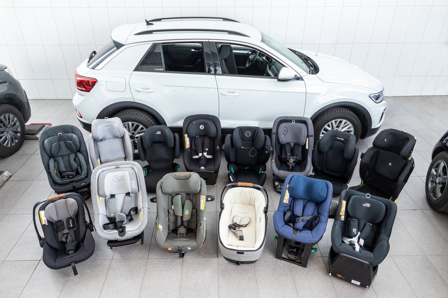 Les sièges auto et coques bébé du test disposés en trois rangées sur un sol carrelé, devant un SUV blanc, vus en plongée