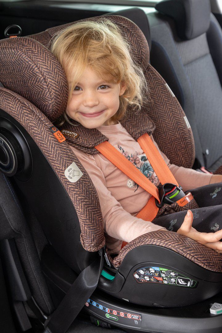 Fillette souriante attachée par un harnais à sangles orange dans un siège auto Cosatto à housse marron à chevrons, installé sur la banquette arrière d'une voiture