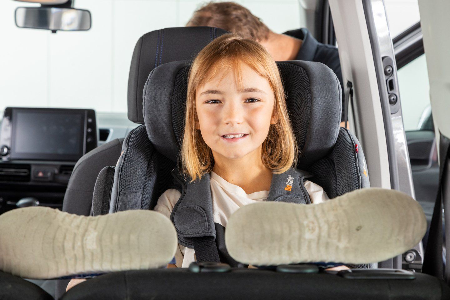 Fillette souriante installée dos à la route dans un siège auto BeSafe gris, les pieds posés en hauteur contre le dossier de la banquette, un adulte visible derrière elle dans la voiture