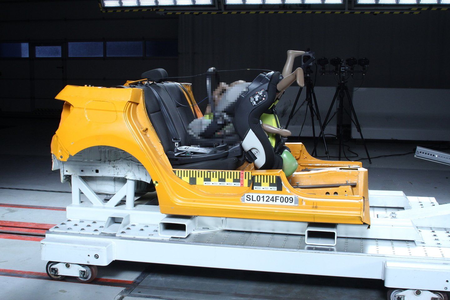 Caisse de voiture jaune sur le chariot d'essai après un crash-test frontal, le siège Peg Perego Viaggio Twist, avec un mannequin enfant, est basculé vers l'avant, jambes du mannequin en l'air, une partie de l'image est pixelisée d'origine