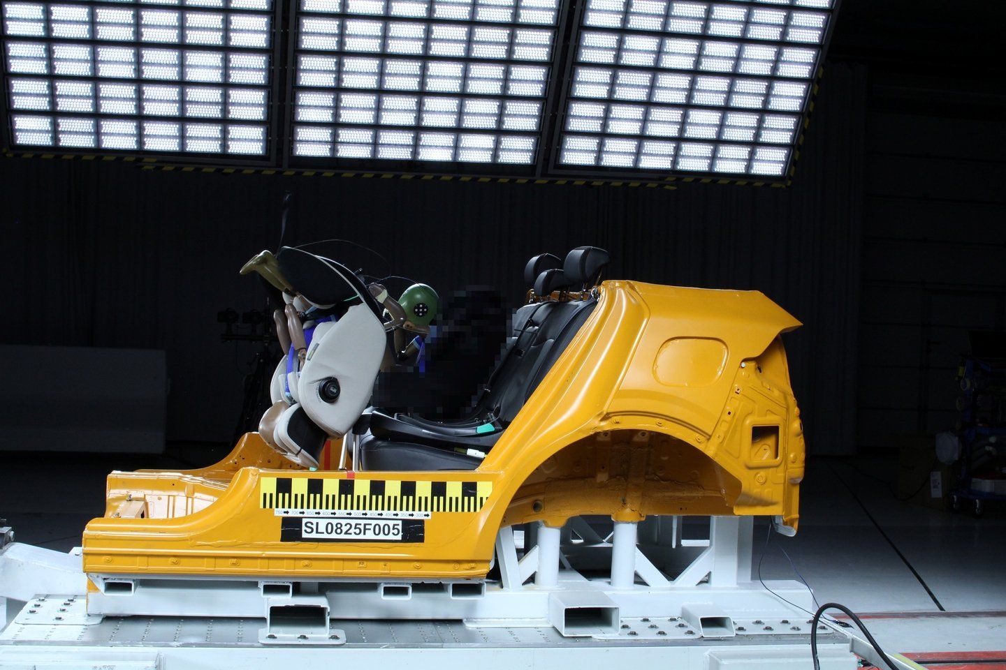 Caisse de voiture jaune fixée sur le chariot d'essai après un crash-test frontal, le siège Chipolino Olympus i-Size, avec un mannequin enfant, a quitté sa place sur la banquette et se retrouve basculé à l'avant de l'habitacle, une zone de l'image est pixelisée d'origine
