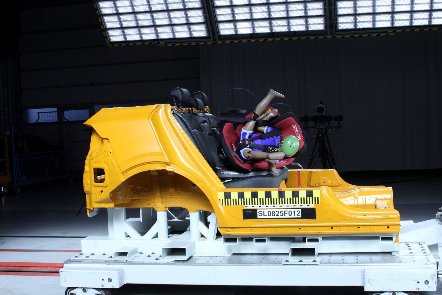 Caisse de voiture jaune sur le chariot d'essai après un crash-test frontal, le siège rouge Reecle 360, avec un mannequin enfant, est renversé et sorti de sa position sur la banquette, une petite zone de l'image est pixelisée d'origine