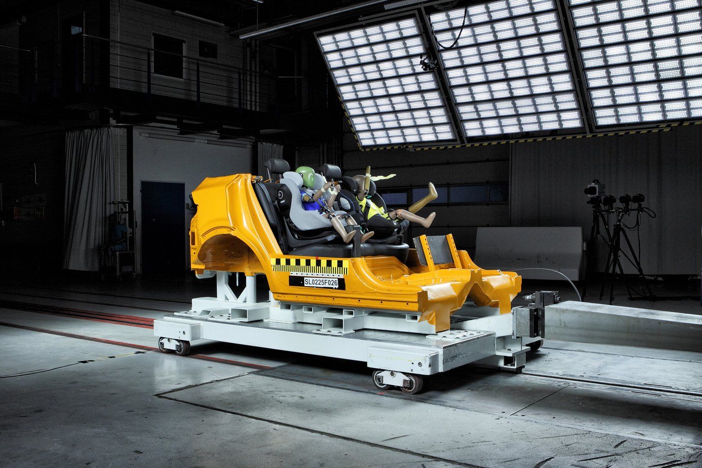 Caisse de voiture jaune fixée sur un chariot d'essai blanc dans un hall de crash-test sombre, sous des panneaux d'éclairage, avec plusieurs mannequins enfants installés dans des sièges auto sur la banquette arrière, avant un choc frontal
