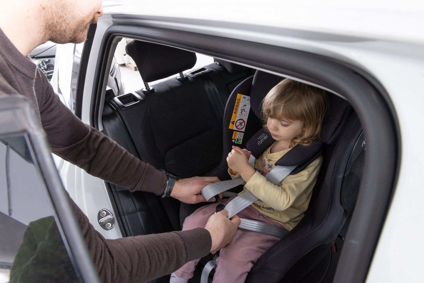 Un adulte, penché par la portière arrière ouverte d'une voiture blanche, boucle la ceinture de sécurité du véhicule sur une fillette assise dans un siège auto Britax Römer noir