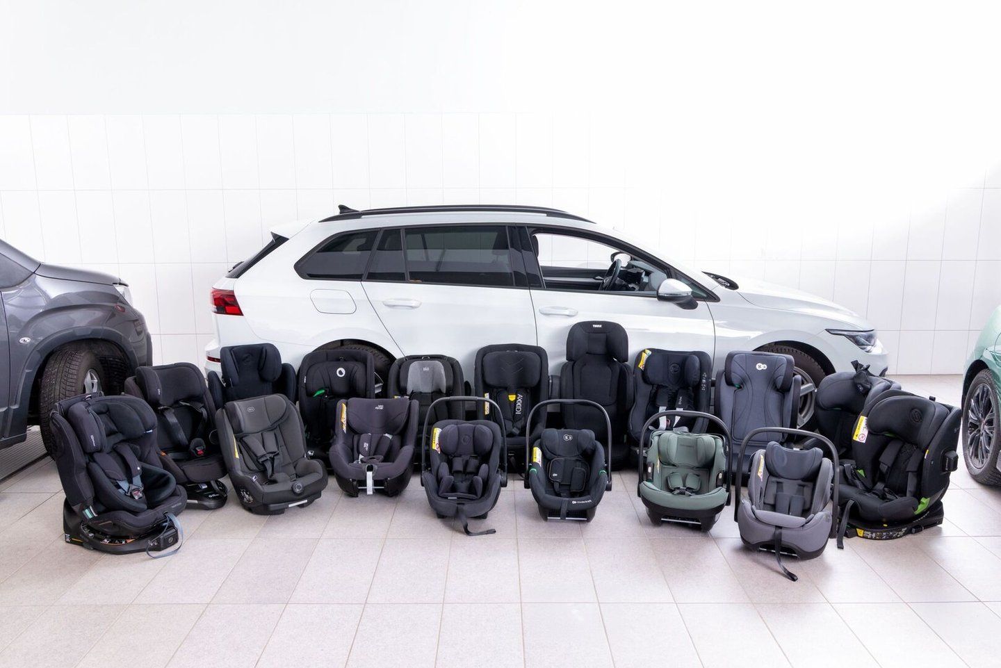 Les sièges auto et coques bébé du test alignés en deux rangées sur un sol carrelé, devant une voiture break blanche, dans un hall d'essai aux murs blancs