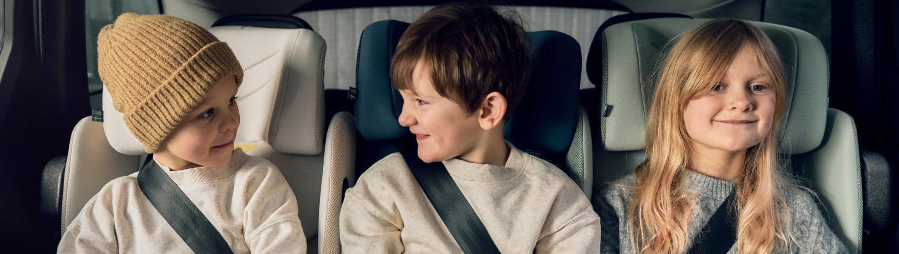 Trois enfants côte à côte à l'arrière d'une voiture, sur des rehausseurs Axkid Up.