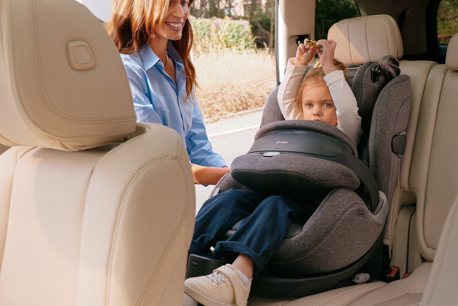 Fillette assise dans le siège auto Cybex Anoris T2 i-Size, retenue par son bouclier à airbag gris, une adulte souriante penchée vers elle depuis la portière arrière