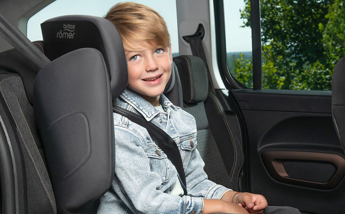 Un garçon blond qui sourit, assis dans le Discovery Plus 2 avec la ceinture de la voiture sur l'épaule