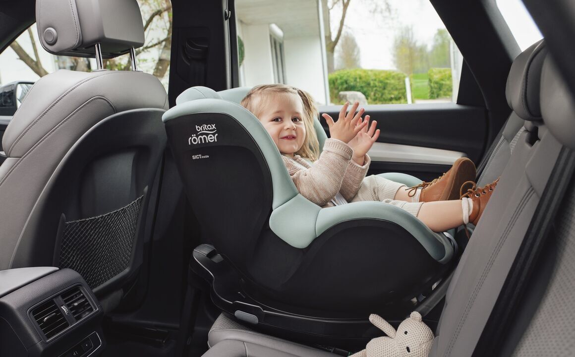 Une petite fille qui applaudit, installée dans un siège vert menthe à l'arrière d'une voiture, face à la portière ouverte