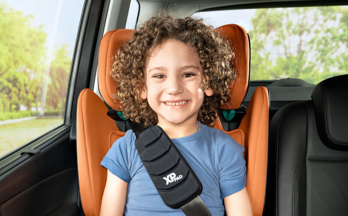 Une fillette aux cheveux bouclés qui sourit, assise dans le Kidfix i-Size orange, la ceinture de la voiture sur l'épaule