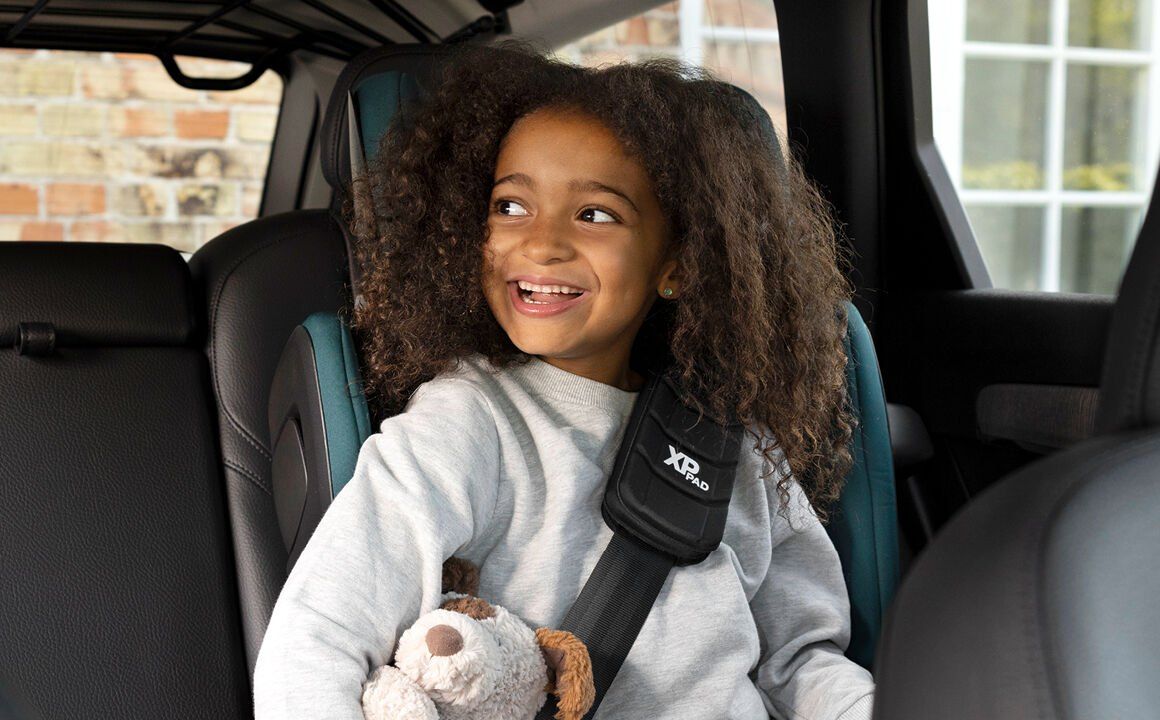 Une fillette aux cheveux bouclés qui rit, assise dans le Kidfix Pro avec un ours en peluche, la ceinture sur l'épaule