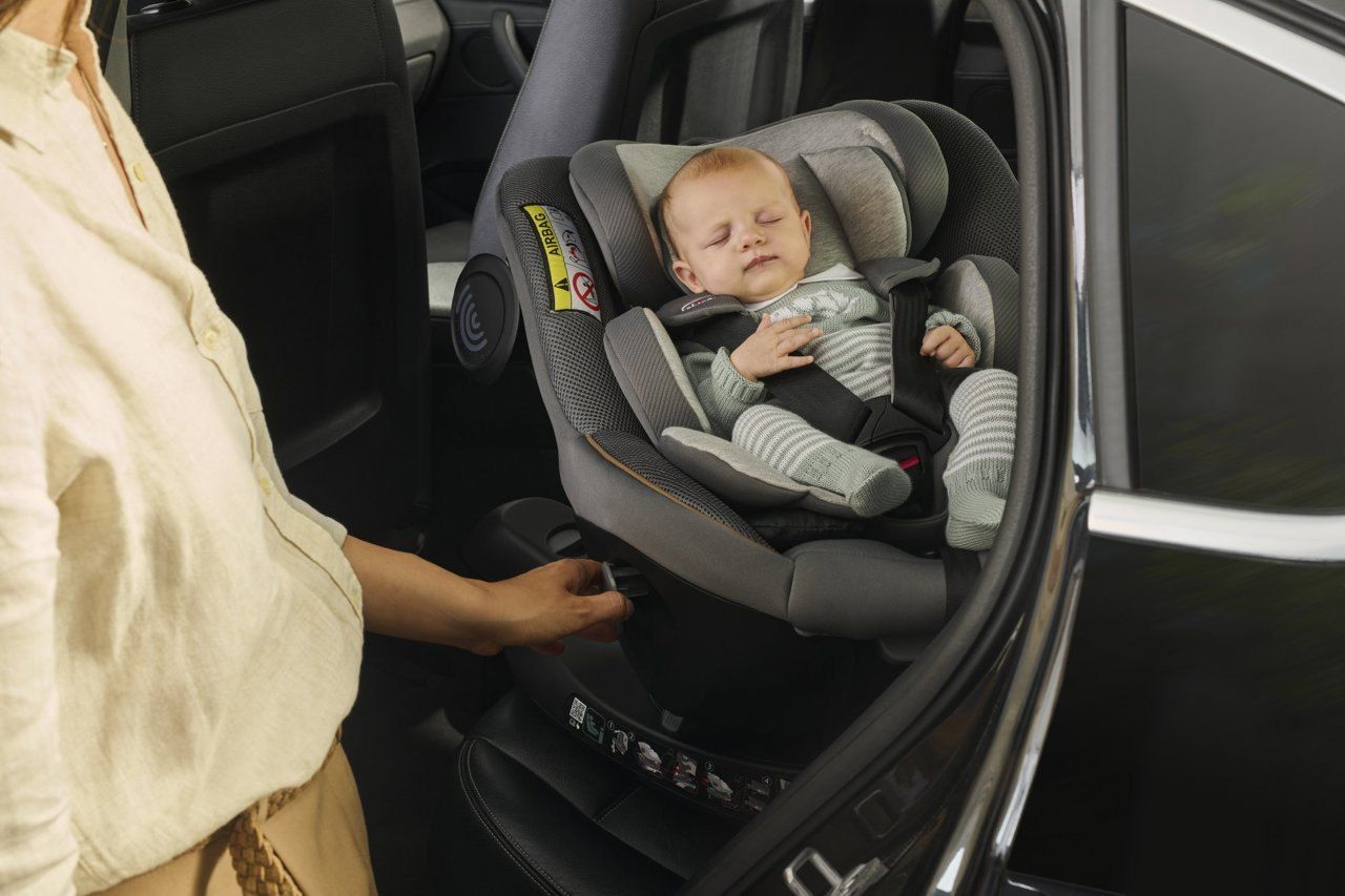 Un bébé endormi dans le siège Chicco Seat105 installé en voiture, la main d'un parent sur la commande de la base.