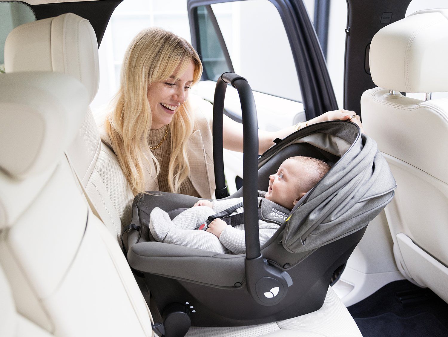 Une adulte souriante penchée vers un bébé attaché dans la coque Joie Sprint grise, installée dos à la route sur la banquette arrière d'une voiture