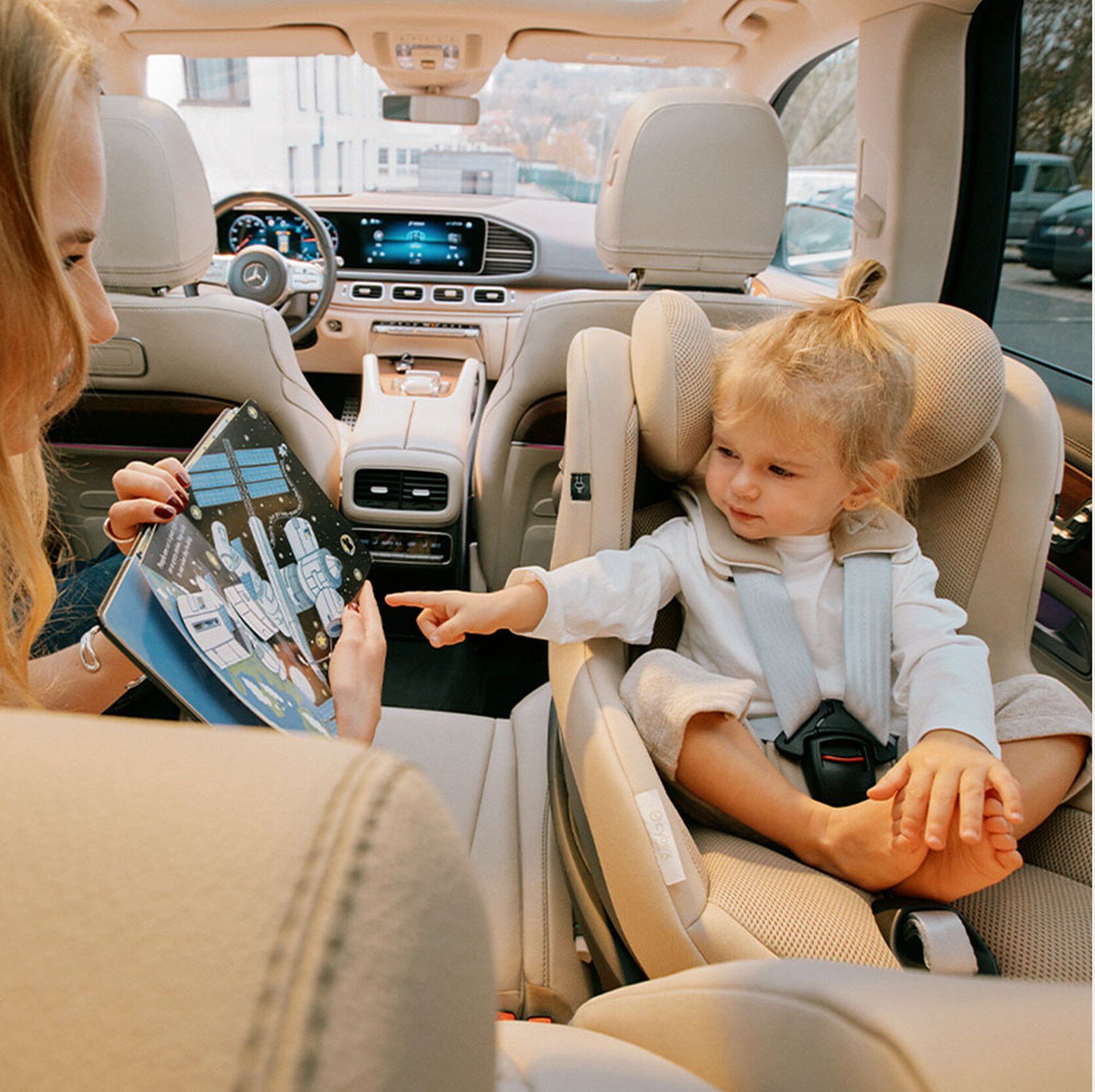 Une petite fille assise dans le siège Cybex Sirona G, face à la route, feuillète un livre avec sa maman.