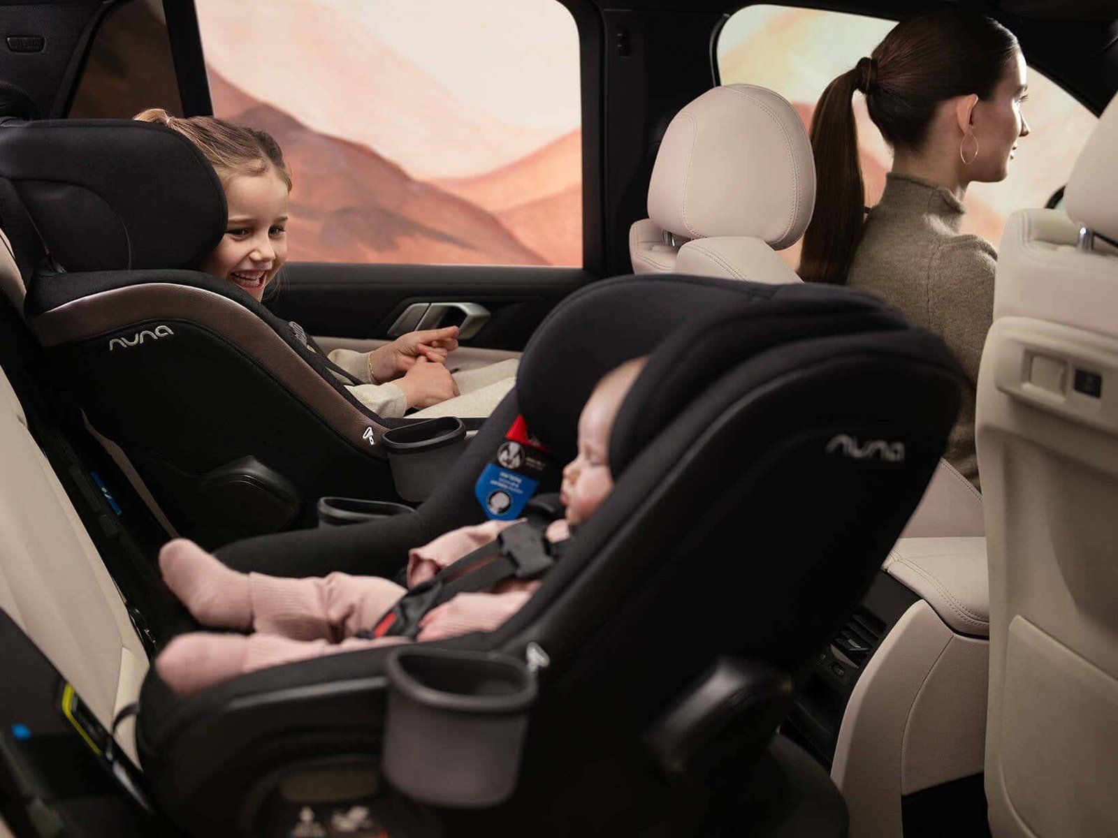 Un bébé dans un siège auto Nuna dos à la route au premier plan, sa grande sœur rieuse dans un autre siège Nuna face à la route, la maman au volant