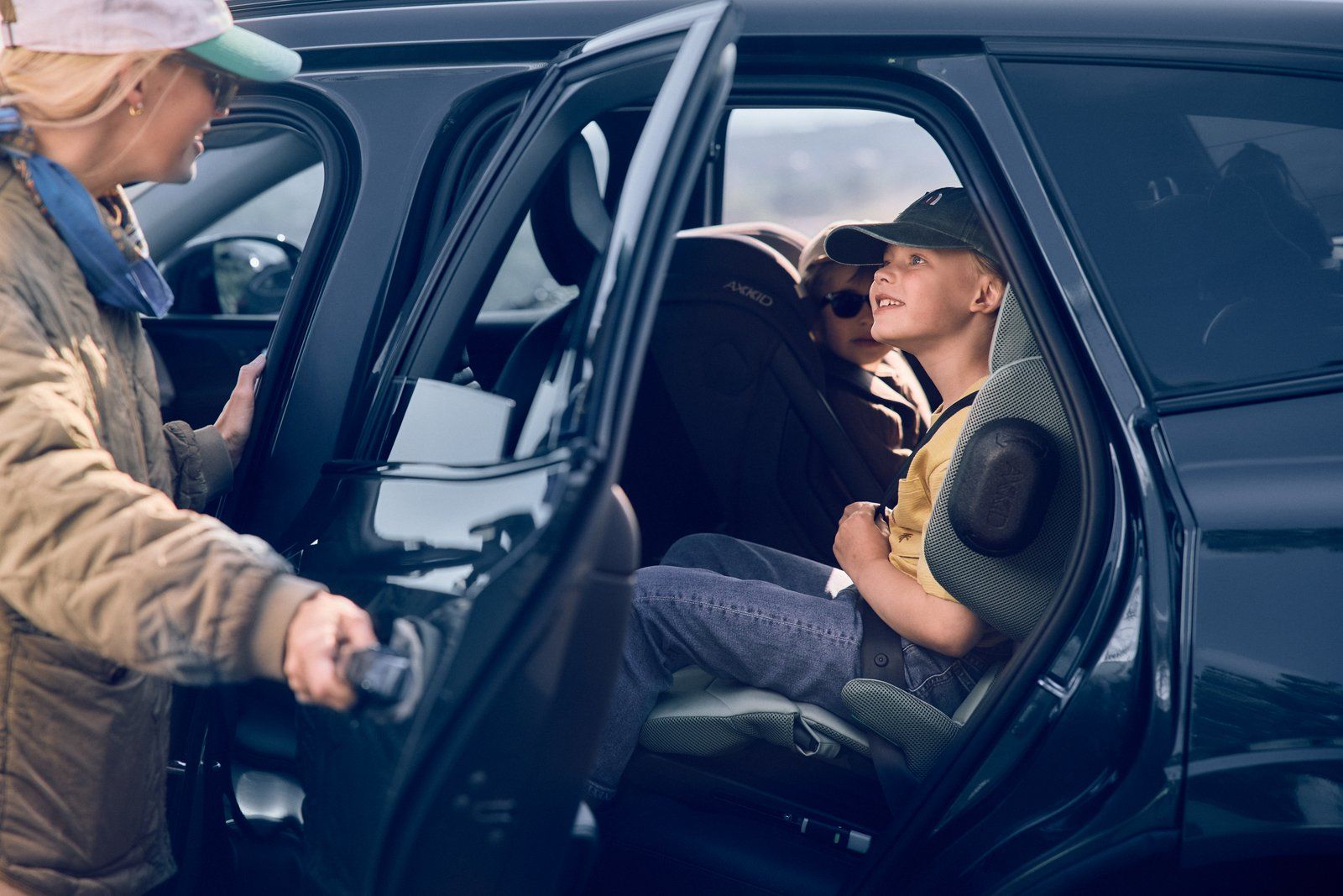 Une femme en casquette ouvre la portière arrière d'une voiture noire, où un garçon attend assis sur son rehausseur Axkid Up à côté d'un autre enfant
