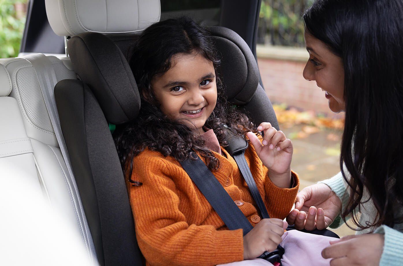Une maman aide une petite fille souriante à boucler son harnais dans un siège Joie i-Irvana en voiture