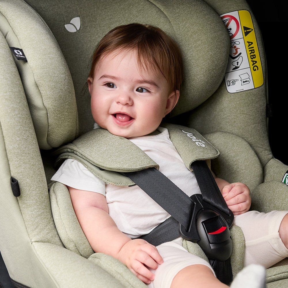 Gros plan sur un bébé souriant, attaché par le harnais dans un Joie i-Spin 360 vert sauge.