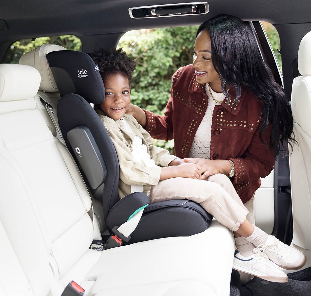 Une maman souriante installe son enfant sur un rehausseur Joie i-Traver à l'arrière d'une voiture.