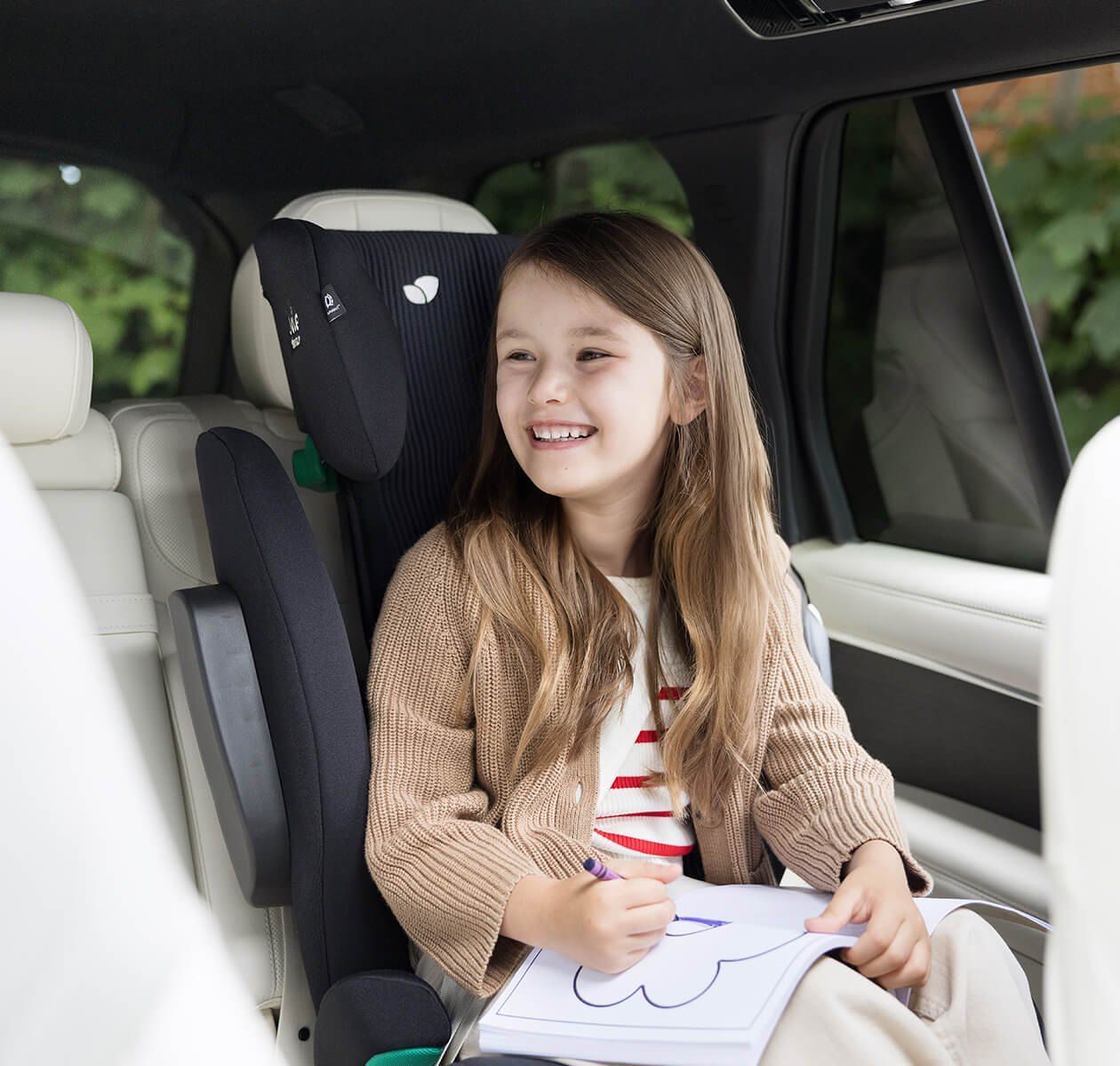 Une fillette souriante dessine, assise sur un rehausseur Joie i-Traver dans une voiture.