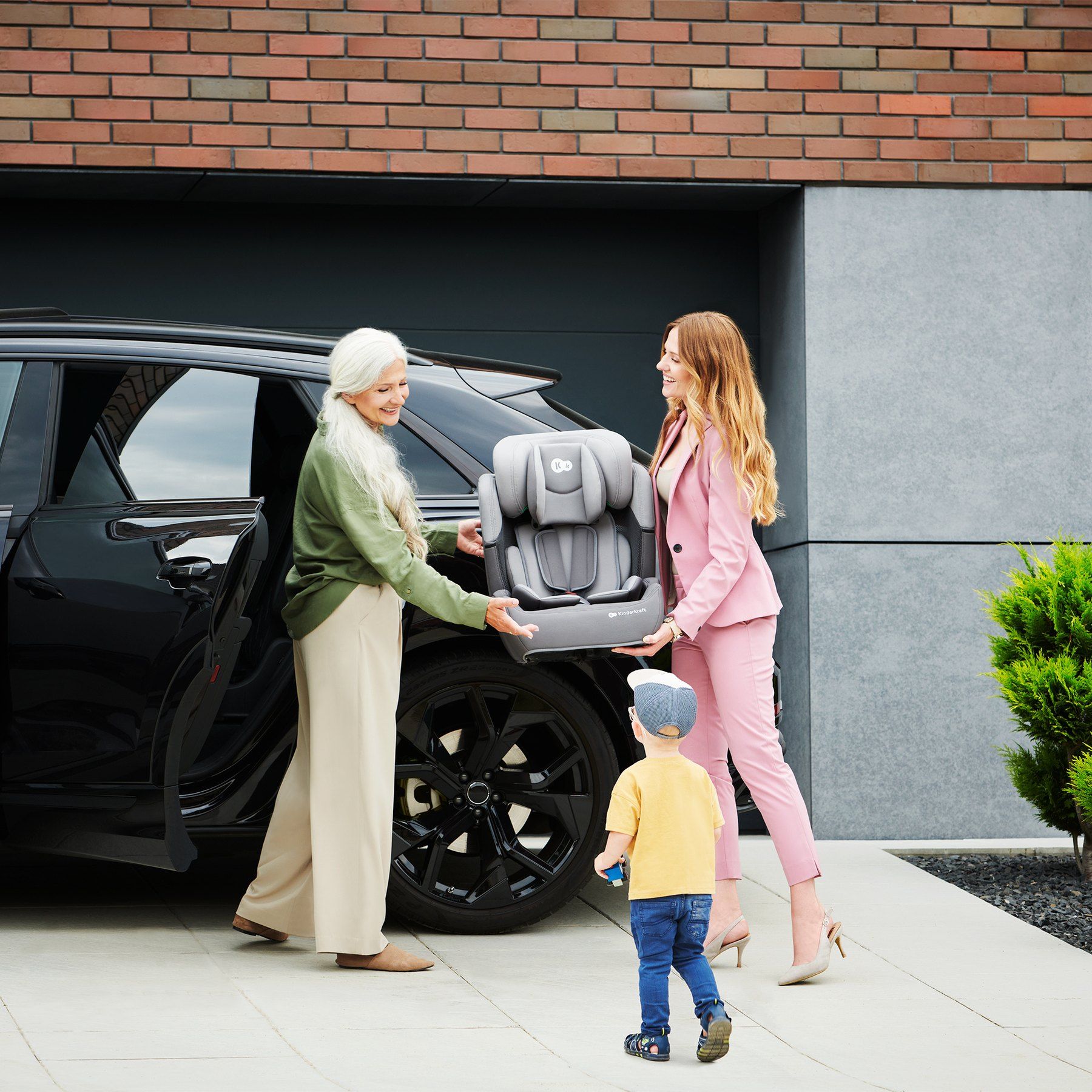 Une grand-mère et sa fille se passent le siège Kinderkraft Comfort Up devant une voiture, un petit garçon à côté d'elles.