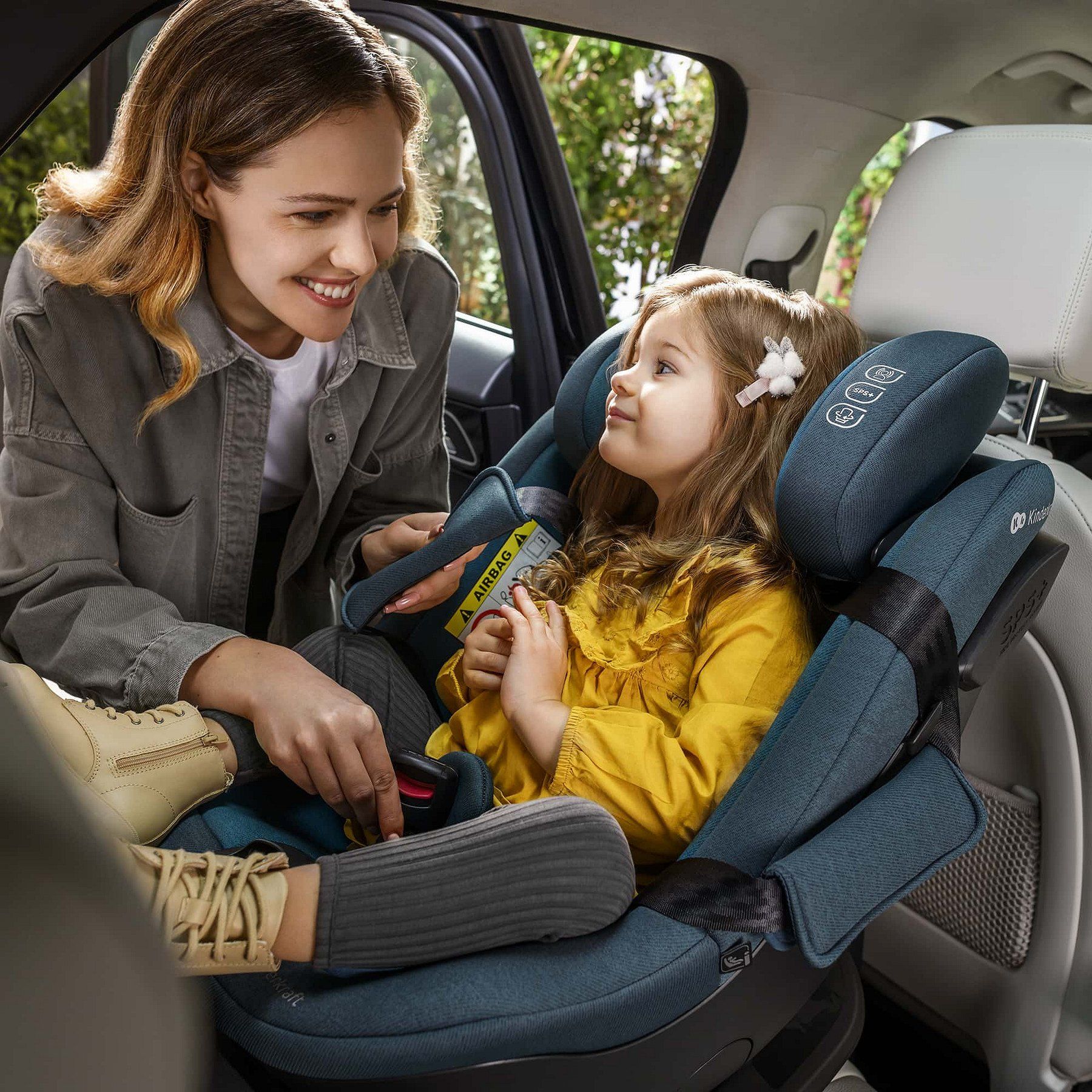 Une maman souriante attache sa petite fille dans le siège Kinderkraft i-Guard, à l'arrière d'une voiture.