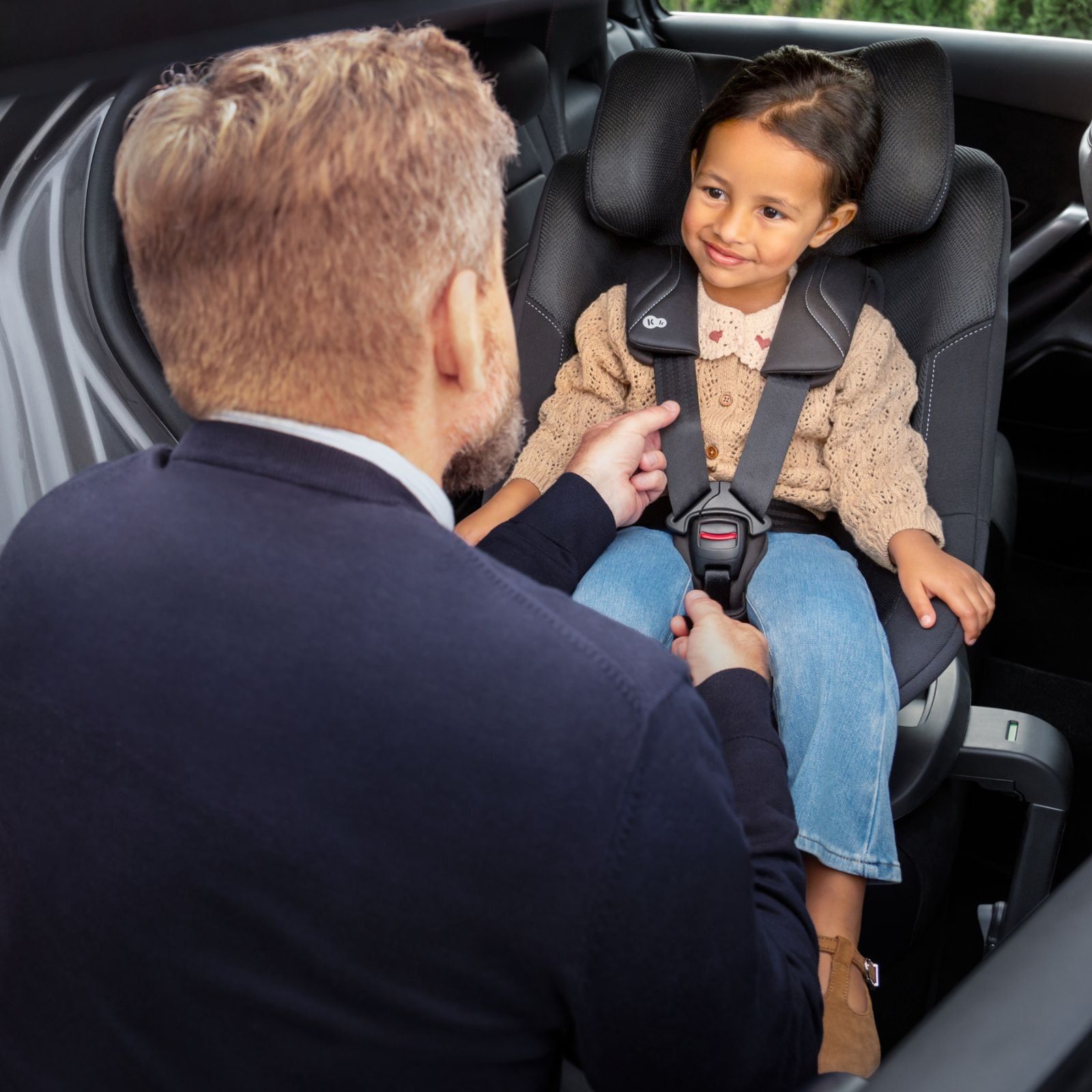 Un papa attache sa petite fille souriante dans le siège Kinderkraft i-Guard Pro, à l'arrière d'une voiture.