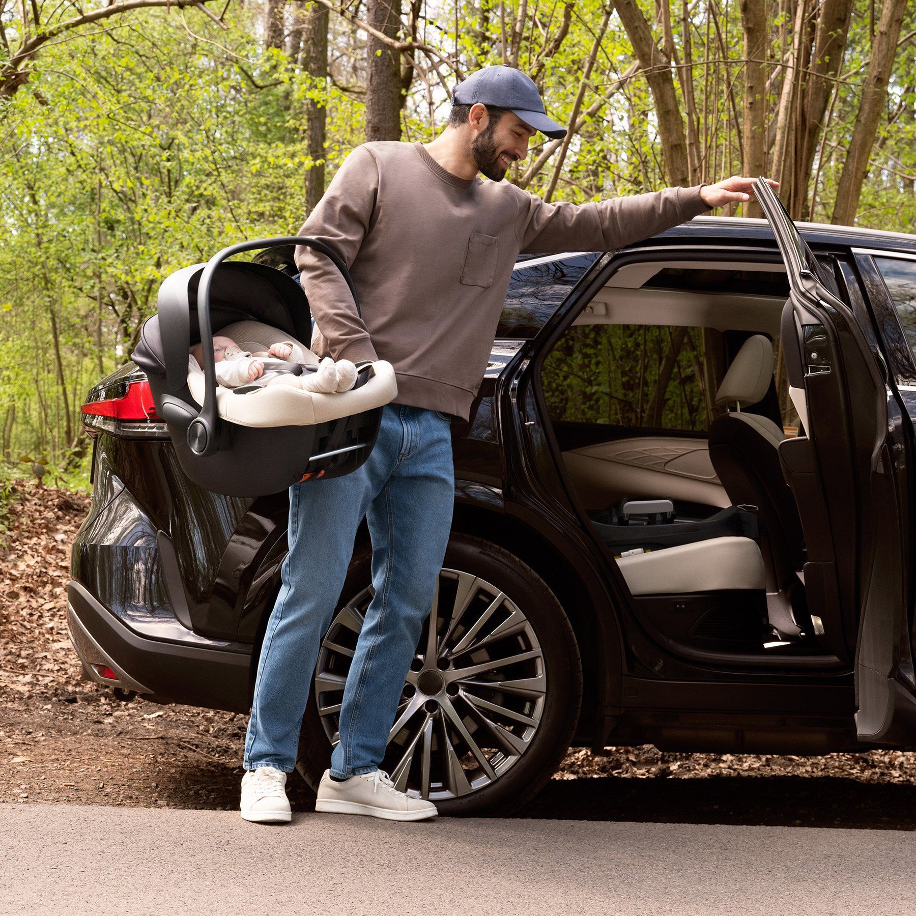 Un papa tient la coque Kinderkraft I-Lite avec son bébé devant la portière ouverte d'une voiture, en forêt.