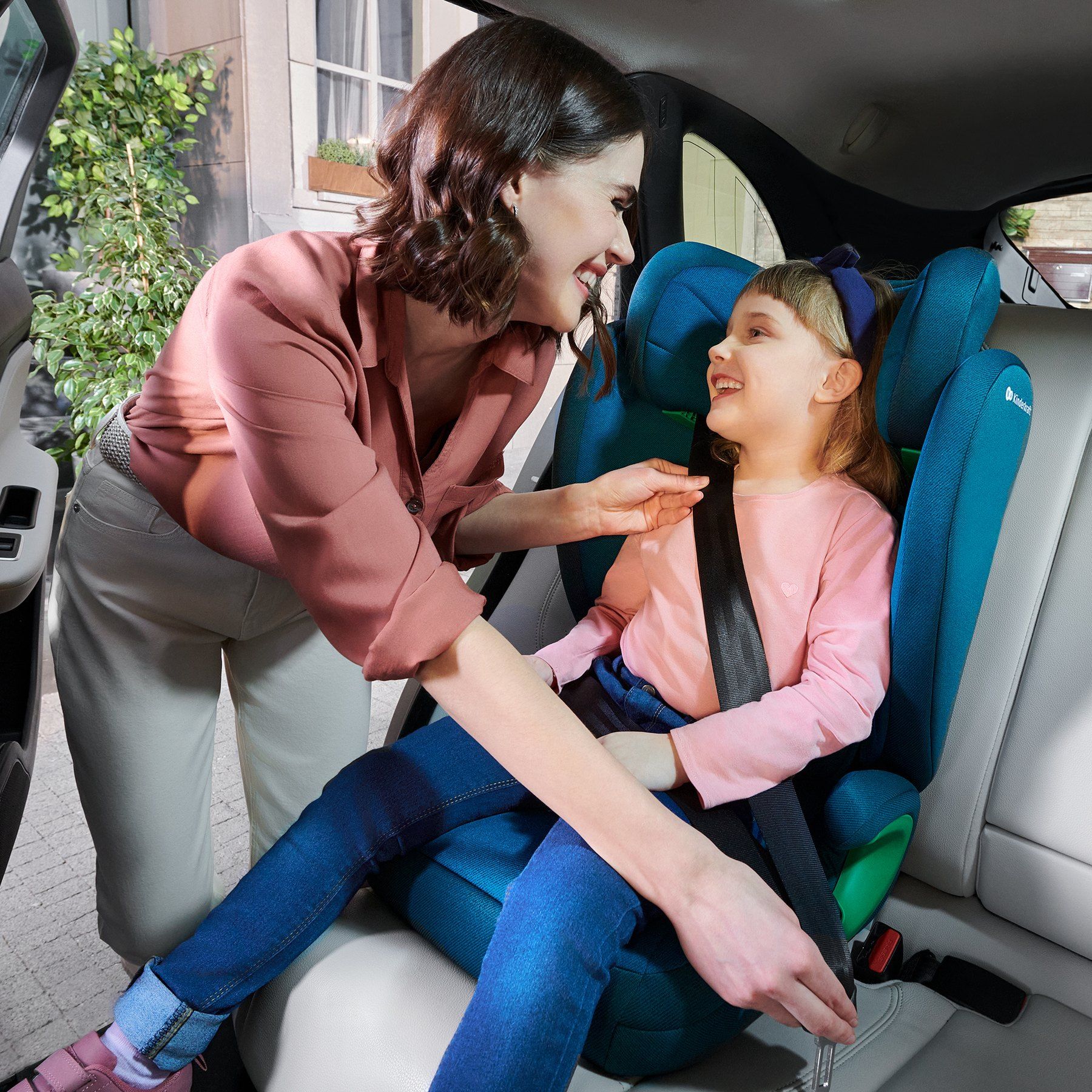 Une maman se penche vers sa fille qui rit, assise sur le rehausseur Kinderkraft Junior Fix 2 i-Size et attachée à la ceinture.