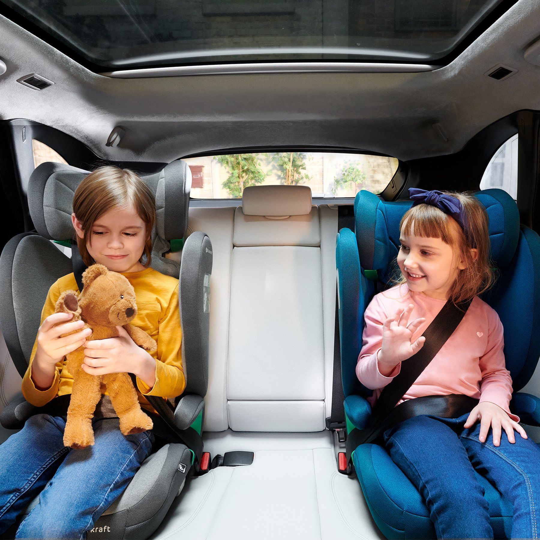 Deux enfants souriants assis côte à côte sur des rehausseurs Kinderkraft Junior Fix 2 i-Size, à l'arrière d'une voiture.