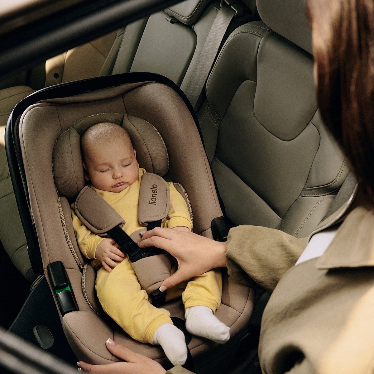 Un bébé endormi en body jaune, attaché par le harnais dans la coque Lionelo Astrid i-Size, à l'arrière d'une voiture