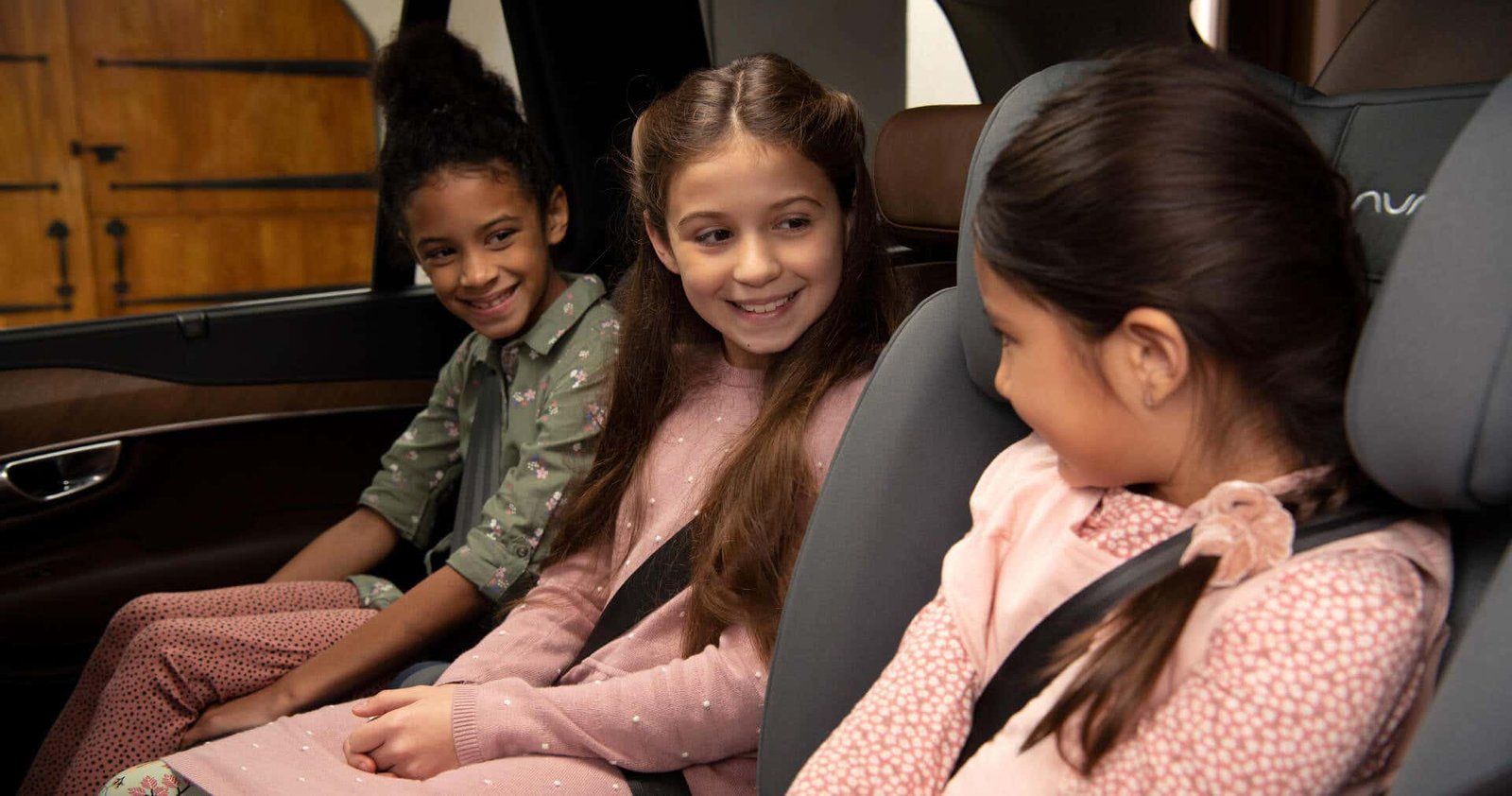 Trois fillettes souriantes assises côte à côte sur des rehausseurs à dossier Nuna, ceinture de la voiture bouclée