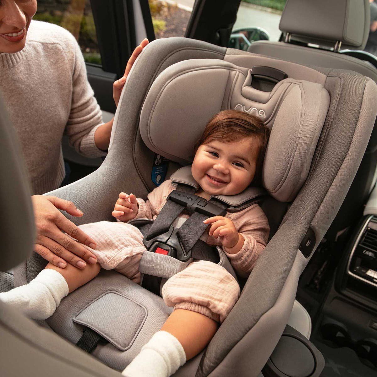 Bébé souriant attaché par le harnais dans un siège auto Nuna gris installé dos à la route, un parent penché vers lui