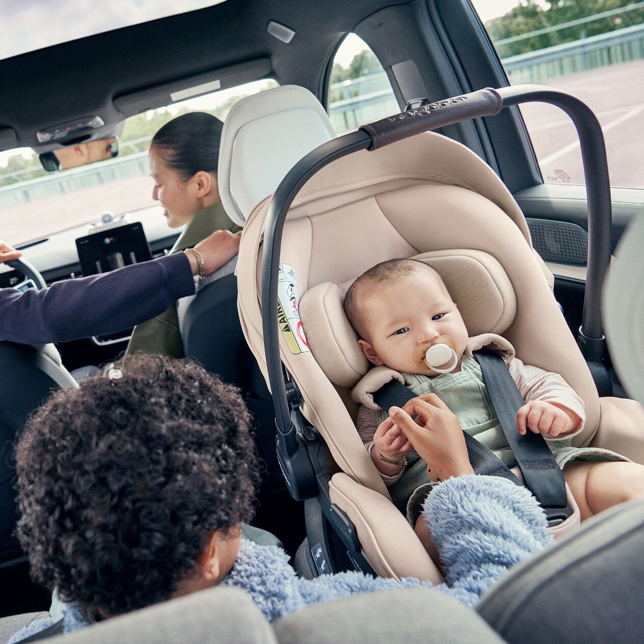Bébé avec sa tétine attaché dans la coque Bugaboo Otter beige, à l'arrière de la voiture, un grand frère à côté de lui et un parent au volant