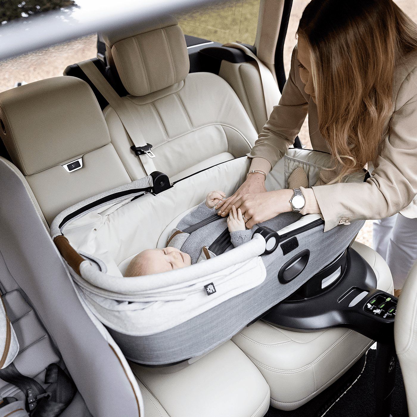 Une adulte attache un nouveau-né allongé dans une nacelle auto Joie grise, posée en travers de la banquette arrière sur sa base ISOFIX