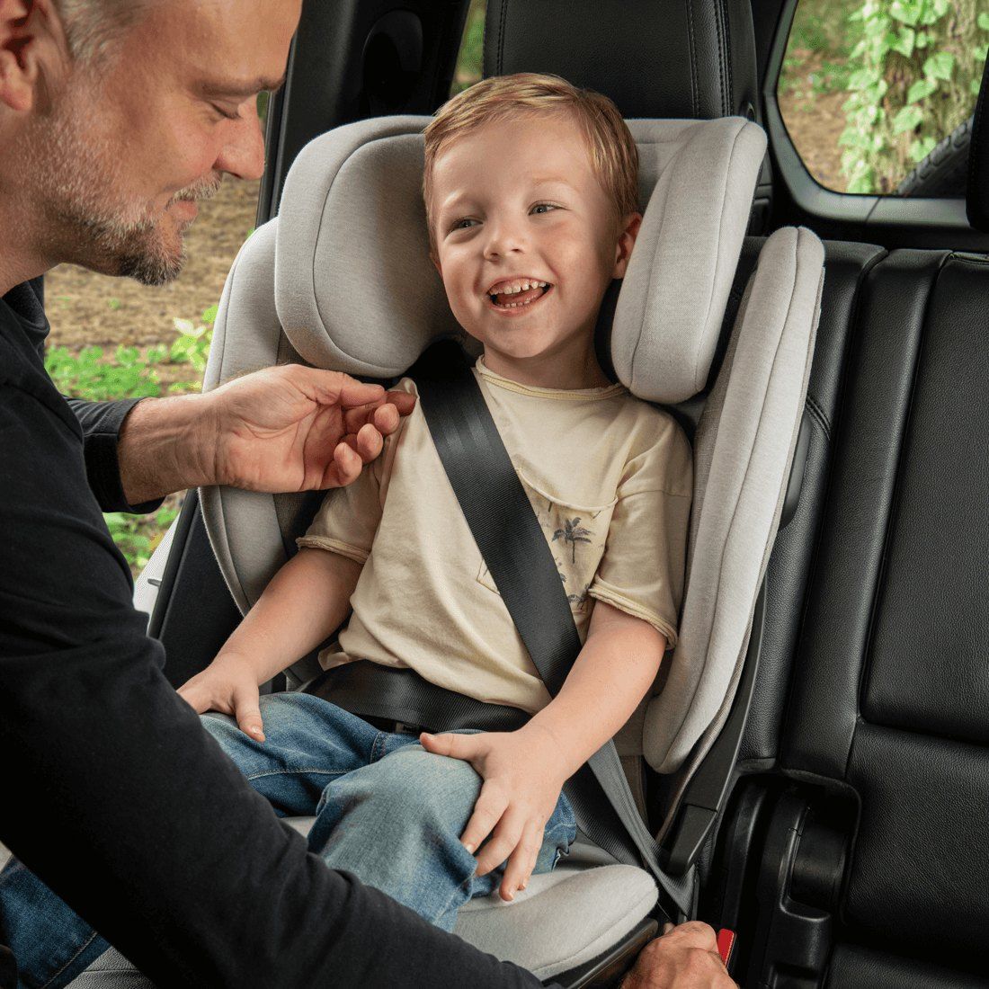 Un papa ajuste la ceinture d'un petit garçon souriant installé sur le rehausseur Nuna Aace LX.