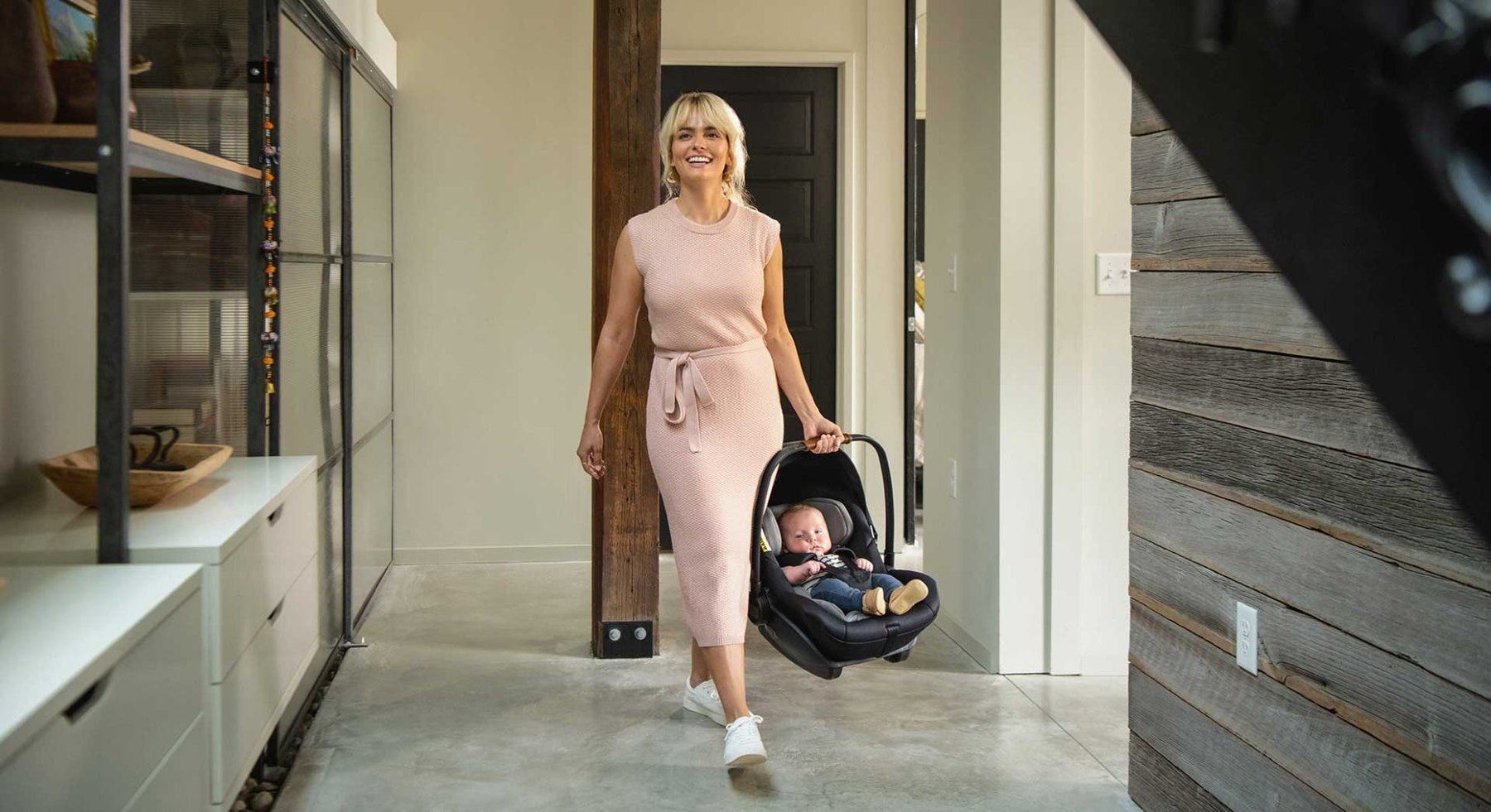 Une maman souriante marche dans une maison en portant la coque Nuna Arra Next avec son bébé.