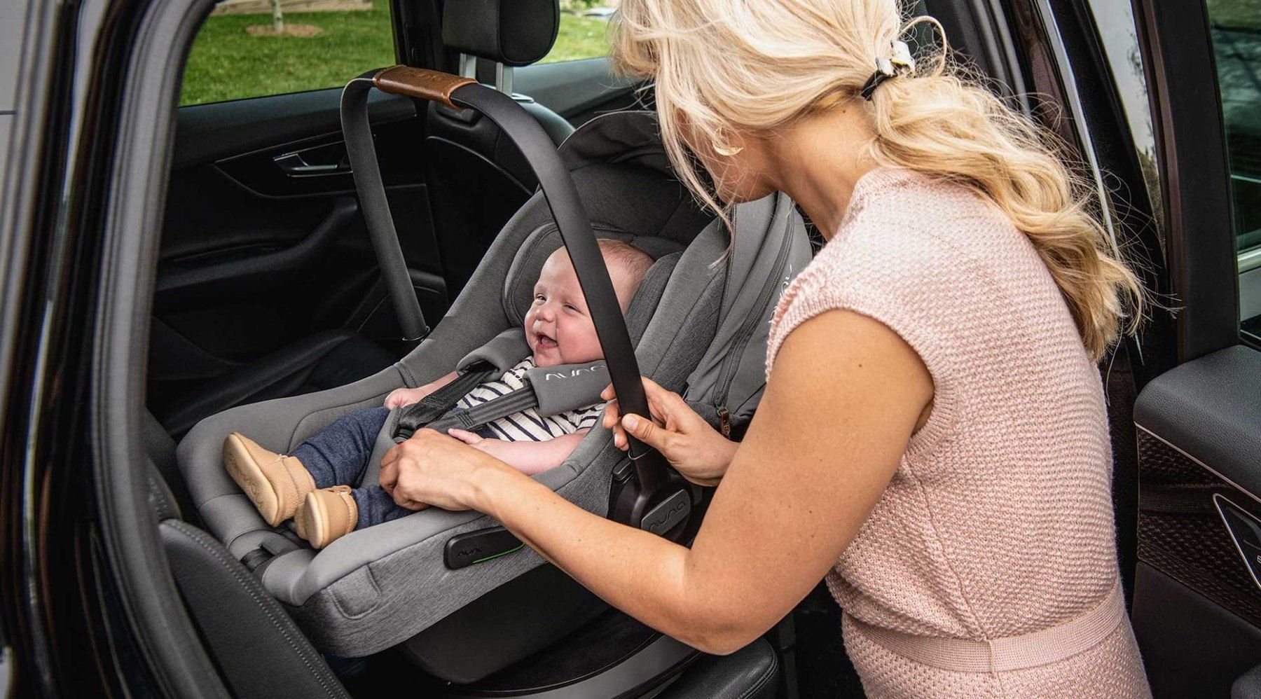Une maman attache son bébé dans la coque Nuna Pipa Next installée dos à la route sur la banquette arrière.