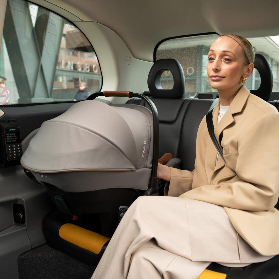 Une femme assise à l'arrière d'un taxi, la coque Nuna Pipa Urbn posée à côté d'elle sur la banquette.