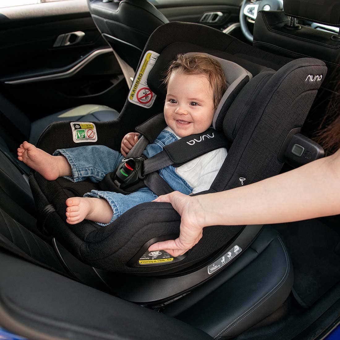 Un bébé souriant installé dans le siège Nuna Todl Next, un parent règle les sangles.