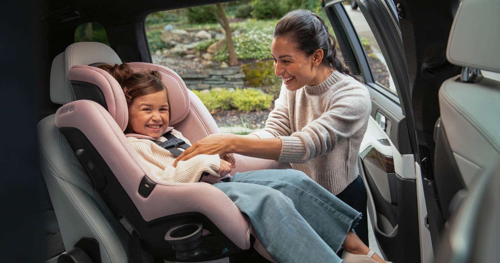 Une maman boucle le harnais de sa fillette d'environ trois ans, rieuse dans un siège auto Nuna rose, portière arrière ouverte