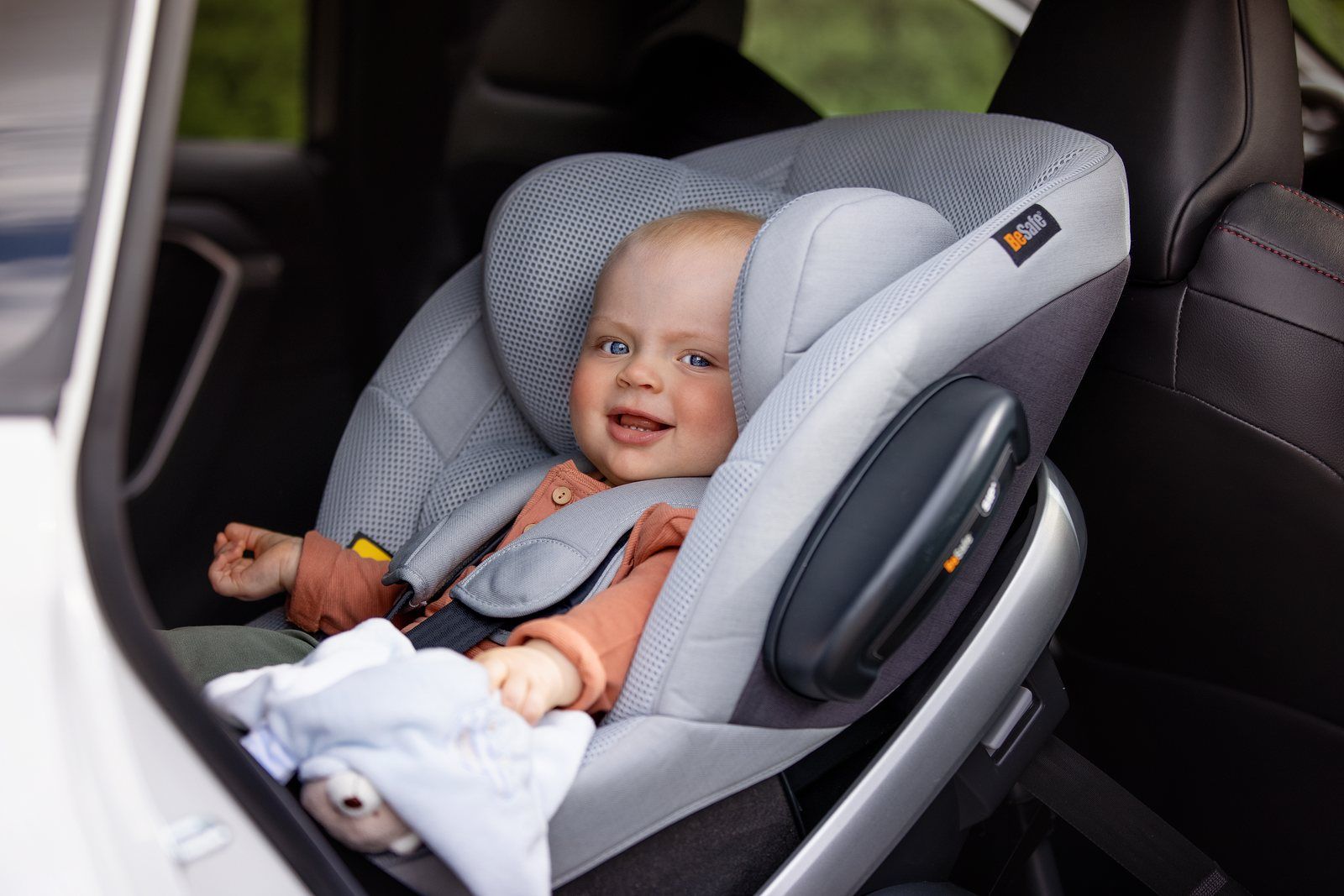 Bébé souriant attaché par le harnais dans le siège auto dos à la route BeSafe Stretch gris, à l'arrière d'une voiture, portière ouverte