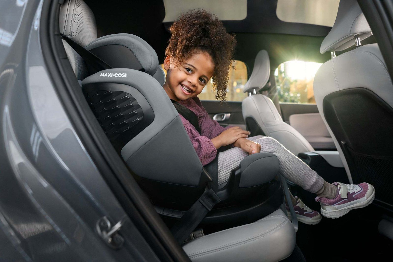 Fillette souriante assise dans le siège auto évolutif Maxi-Cosi Emerald 360 S gris, en mode rehausseur avec la ceinture de la voiture, portière arrière ouverte