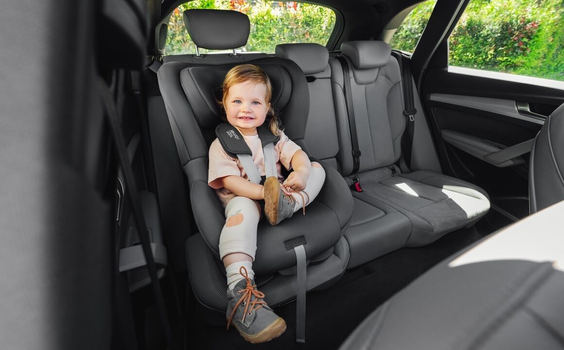 Une petite fille souriante attachée par un harnais dans un siège auto Britax Römer installé face à la route sur la banquette arrière