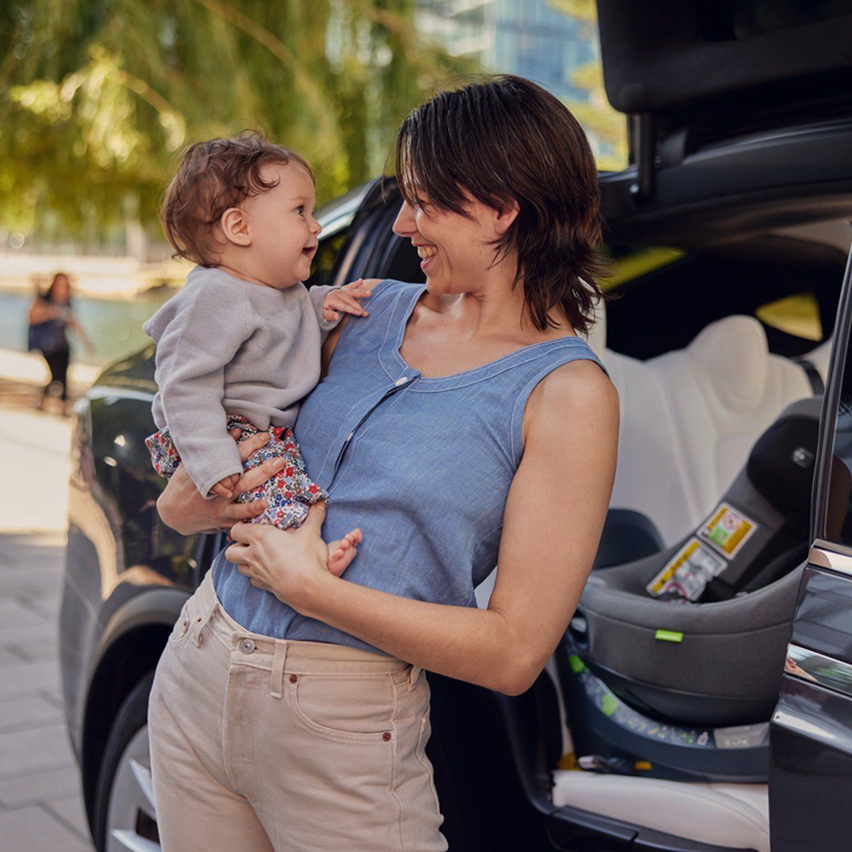 Une maman porte son bébé devant la portière ouverte, avec un Swandoo Curie installé dans la voiture.