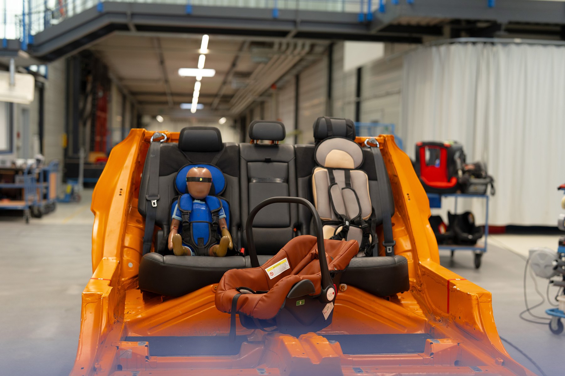Une coque bébé marron et deux sièges à harnais achetés en ligne, installés sur la banquette d'une caisse de crash-test, un mannequin dans l'un d'eux